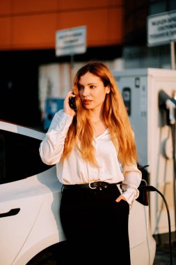 Kızıl saçlı bir iş kadını gün batımında elektrikli arabayı şarj ediyor.