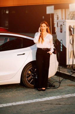 Kızıl saçlı bir iş kadını gün batımında şarj istasyonunda elektrikli araba yüklüyor.