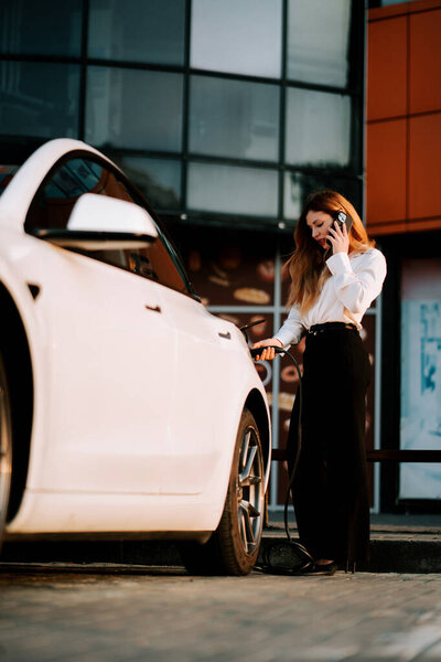 Деловая женщина с рыжими волосами заряжает электрический автомобиль на станции во время разговора по телефону