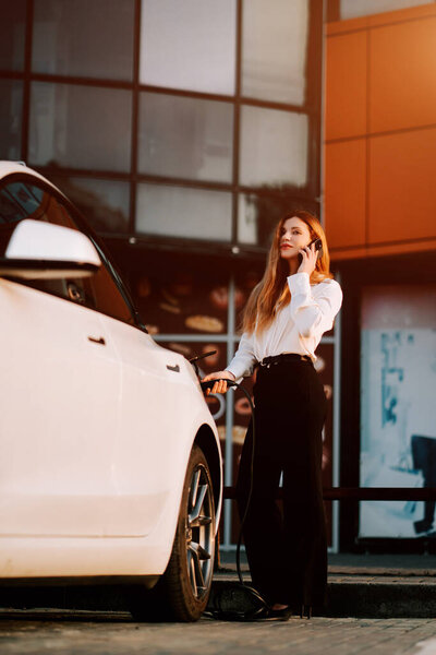 Деловая женщина с рыжими волосами заряжает свой электромобиль на зарядной станции в городской местности