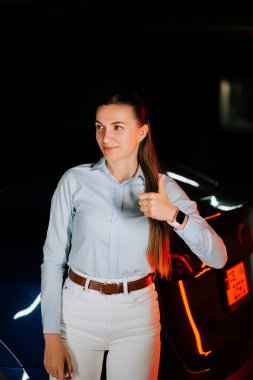 EV 'yi yeraltı otoparkında şarj eden kadın, tatmin olmak için başparmağını kaldırıyor.