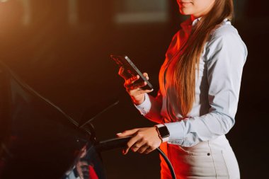 Kadın şehir ortamında akşam saatlerinde neon ışıklarının altında elektrikli araba şarj ediyor.