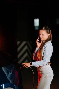 Kadın, canlı neon ışıkların altında telefonla konuşurken elektrikli arabayı şarj ediyor.