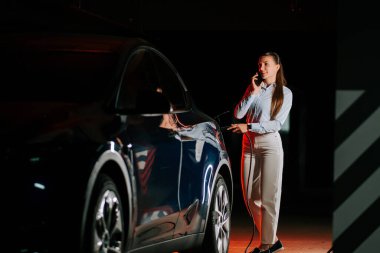 Kadın, neon ışıklı garajda telefonla konuşurken elektrikli arabayı şarj ediyor.