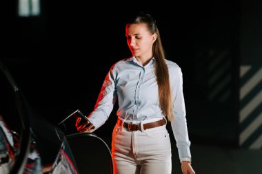 Kadın, şehrin gece lokasyonunda canlı neon ışıkla elektrikli arabayı şarj ediyor.