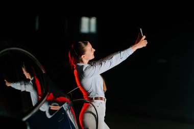 Gece şehir ortamında neon ışıklarının altında elektrikli araba yükleyen bir kadın.