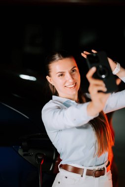 Kadın selfie çekerken neon ışıklı ortamda elektrikli arabayı şarj ediyor.