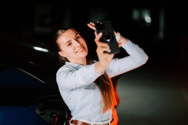 Elektrikli arabayı neon ışıkta şarj eden kadın gece selfie çekerken gülümsüyor.