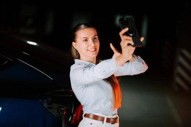 Kadın gece neon ışıklarının altında elektrikli araba şarj ederken akıllı telefon kullanıyor.