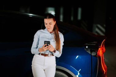 Kadın, gece vakti neon ışıklı şarj istasyonunda elektrikli araç şarj ediyor.