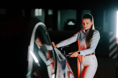 Kadın şehir şarj istasyonunda gece saatlerinde neon ışıkta elektrikli araba şarj ediyor.