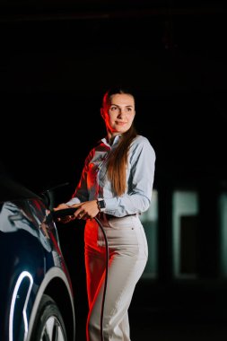 Modern park alanında kadın elektrikli aracı neon ışıkların altında şarj ediyor.