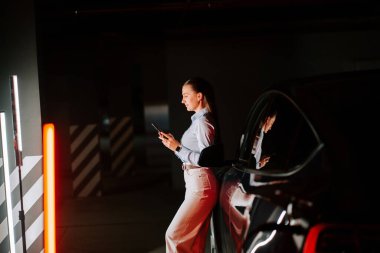 İş kadını, yeraltı otoparkında telefon kullanırken arabayı şarj ediyor.