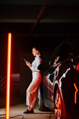 İş kadını, yeraltı otoparkında akıllı telefon kullanırken elektrikli arabayı şarj ediyor.