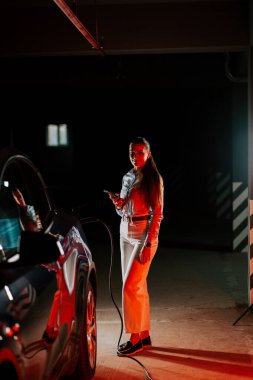 Yeraltındaki otoparkta elektrikli aracı şarj ederken telefonu kullanan bir iş kadını.