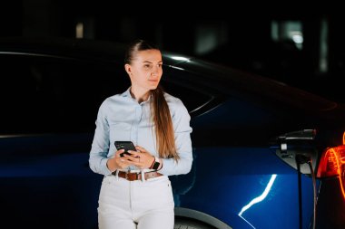 İş kadını, yeraltı otoparkında akıllı telefon kullanırken elektrikli aracı şarj ediyor.