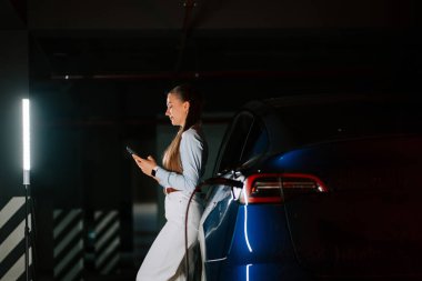 Yeraltı otoparkında akıllı telefon kullanan bir iş kadını elektrikli aracı şarj ediyor.
