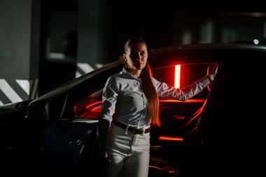 Gece vakti şehir ortamında uzun saçlı genç bir kadın arabanın yanında duruyor.