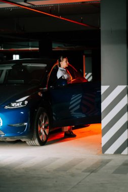 Yeraltı otoparkında sıcak ışıklandırmayla mavi arabadan inen kadın.