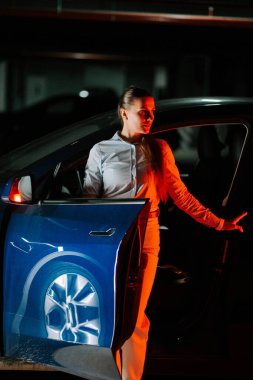 Gece vakti mavi arabadan çıkan kadın çarpıcı ışıklandırma efektleri gösteriyor.