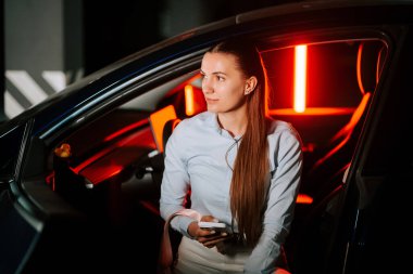 Kadın, akşam saatlerinde modern otoparkta arabasında oturmuş elinde telefon tutuyor.
