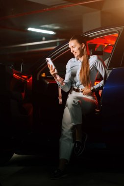 Otoparkta loş ışıkta otururken akıllı telefon kullanan genç bir kadın.