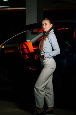 Gecenin bir yarısı loş bir otoparkta arabanın yanında güven içinde poz veren bir kadın.