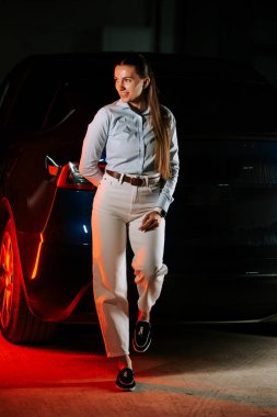 Şık bir kadın loş bir garaj ortamında modern bir arabanın yanında güven içinde poz veriyor.