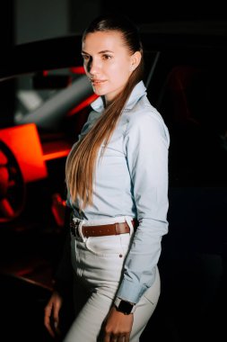 Açık mavi gömlekli genç kadın loş ışıklı bir arabanın yanında güven içinde poz veriyor.