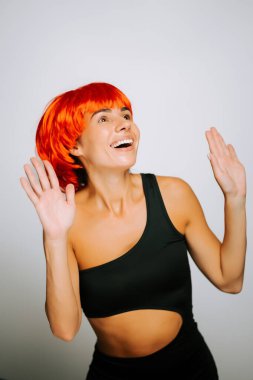 Parlak kırmızı peruklu bir kadın basit arka planda siyah kıyafetiyle neşeyle dans ediyor.