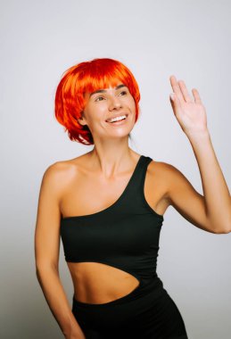 Parlak turuncu peruklu, gülümseyen bir kadın. İçerde siyah elbisesiyle neşeyle el sallıyor.