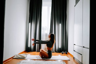 Gündüz vakti huzurlu bir evde yoga yapan genç bir kadın.