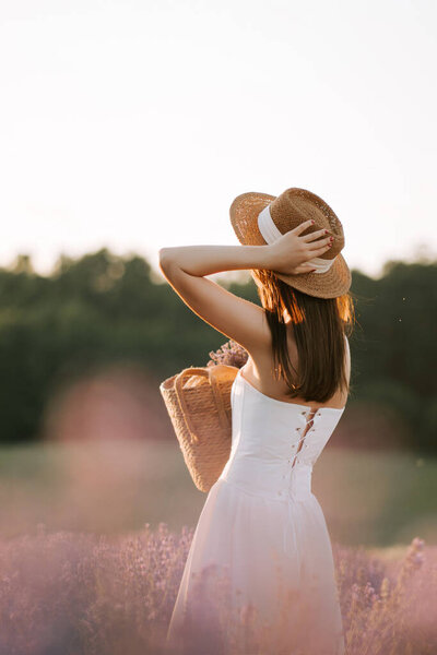Woman in white dress with straw hat enjoying lavender field at sunset