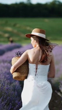 Beyaz elbiseli kadın lavanta tarlasında elinde sepetle yürüyor.