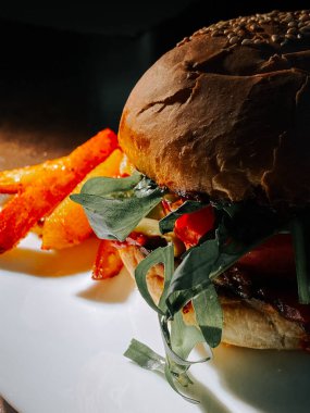Yumuşak ışıklandırılmış beyaz bir tabakta servis edilen taze soslu sulu burger ve kızarmış patates.