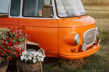 Renkli çiçeklerle dolu, güneşli bir tarlada, ekicilerin yanına park edilmiş turuncu bir minibüs.