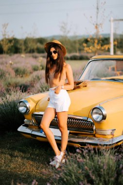 Yaz kıyafetli genç bir kadın gündüz vakti lavanta tarlasında sarı bir arabanın yanında poz veriyor.