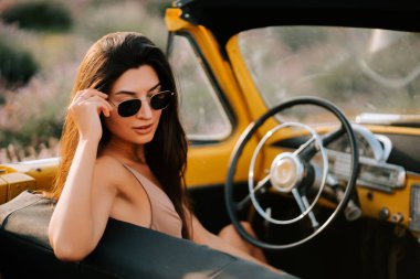 Güneş gözlüklü çarpıcı kadın gün batımında manzaralı bir yerde klasik bir üstü açık arabadan hoşlanıyor.