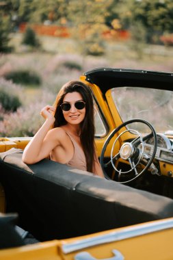 Güneşli bir günde, sarı, üstü açık bir arabada gülümseyen genç bir kadın.