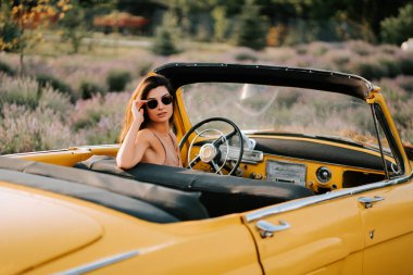 Güneş gözlüklü genç bir kadın güneşli bir günde üstü açık sarı bir arabayla gezintiye çıkıyor.