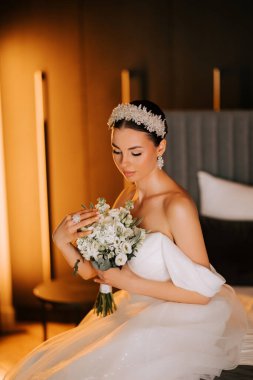 Şık bir gelinlik içinde, seremoniden önce yumuşak bir şekilde aydınlatılmış iç mekanda bir buket tutuyor.