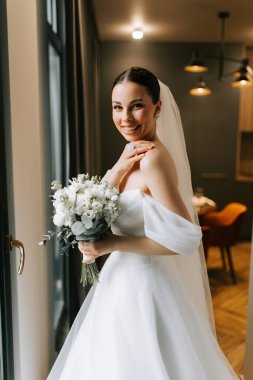 Gelin, düğün töreninden önce pencerenin yanında gülümserken buket tutuyor.