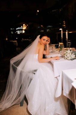 Şık elbiseli gelin akşam resepsiyonunda mum ışığında masada oturuyor.