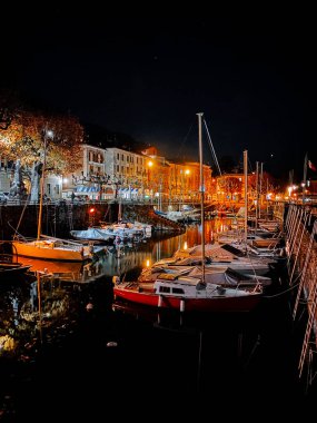 Gece liman manzarası, aydınlatılmış tekneler ve kıyı kasabasındaki deniz kıyısındaki binalar.