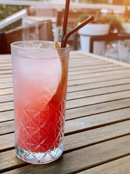 Gün batımında ahşap bir masada buzlu uzun bir bardakta servis edilen renkli bir kokteyl.