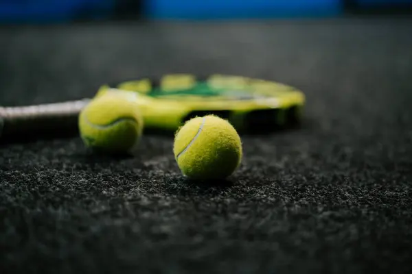 Tenis topları ve raket bir spor tesisinde dokulu bir yüzey üzerinde dinleniyor.