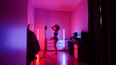 Kadın renkli neon ışıklı odada dans ederken telefonundan canlı yayın yapıyor.