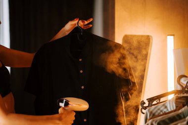 Modern ev ortamında siyah gömlek ütülemek için vapur kullanan bir kadın.