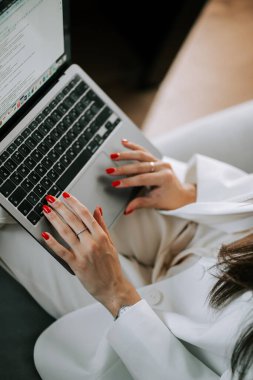 Modern bir iç mekanda koltukta otururken dizüstü bilgisayarda yazan profesyonel bir kadın.