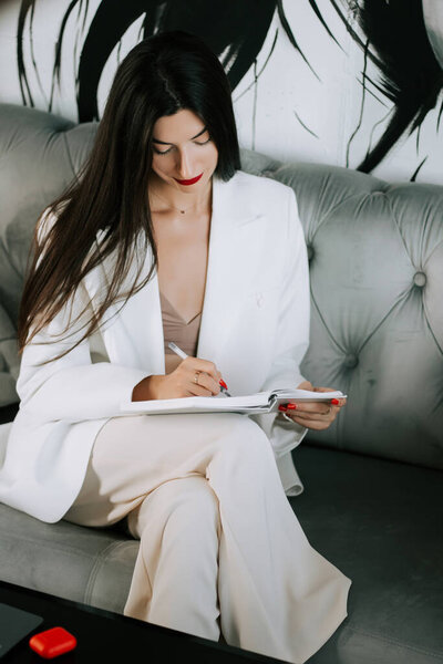 Woman in white suit writing notes while sitting on a stylish couch in a modern interior